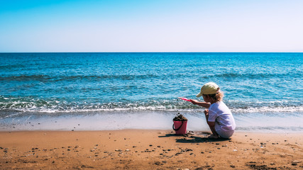 A child plays with a pink plastic bucket and shovel in the sand on a beach, close to the sea....