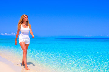 Gorgeous blond woman with white clothes is walking in the water on the sandy beach