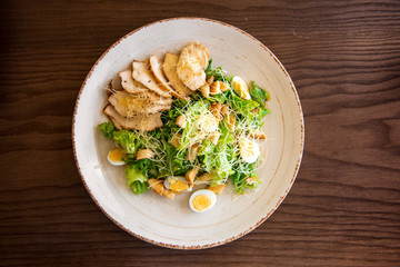 Fresh summer salad Ceasar with eggs, lettuce, chicken and parmesan cheese on white plate on wooden background; healthy concept; top flatlay view