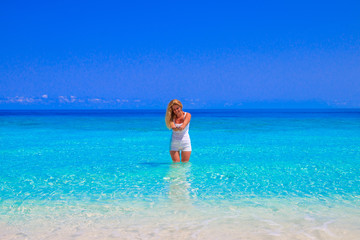 Gorgeous blond woman with white clothes is standing in the water at the sandy beach