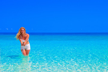 Fototapeta na wymiar Gorgeous blond woman with white clothes is standing in the water at the sandy beach