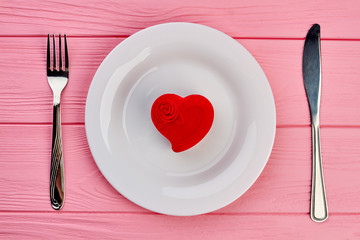 Saint Valentines Day table setting. White plate, red velvet box with present and cutlery on pink wooden background, top view. Valentines Day concept.
