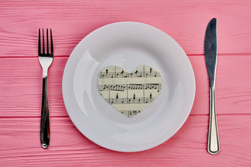 Table setting with cutlery and paper heart. Paper heart with music notes, plate, silver fork and knife, top view.