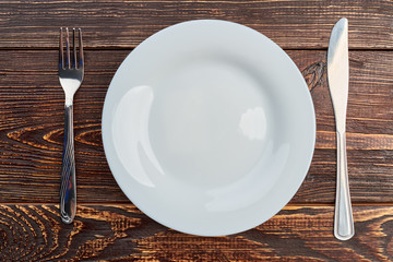 Table setting with plate, fork and knife. White empty plate, silver fork and knife on dark wooden background, top view.