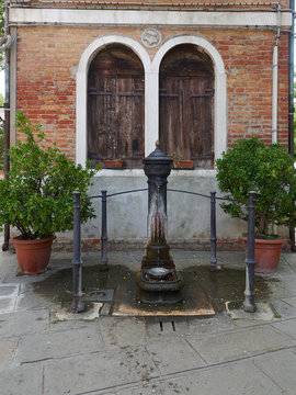 Decorative fountain with a bowl of water for dogs, flanked by two flower pots, in front of a house in Murano, Venice, Italy