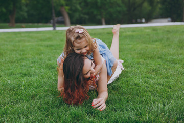 Fototapeta premium Joyful woman in light dress and little cute child baby girl lie on green grass in park rest play and have fun. Mother, little kid daughter. Mother's Day, love family, parenthood, childhood concept.
