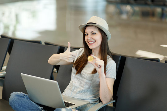 Young smiling traveler tourist woman in hat sitting, working on laptop, holding bitcoin, showing thumb up, waiting in lobby hall at airport. Passenger traveling abroad on weekend. Air flight concept.