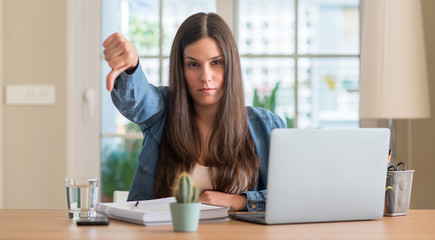 Young student woman studying at home with angry face, negative sign showing dislike with thumbs...