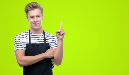 Young handsome blond man wearing apron very happy pointing with hand and finger to the side