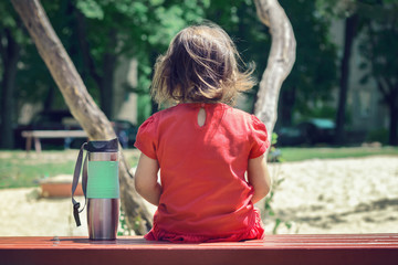 A little upset girl in a red dress sits on a bench alone