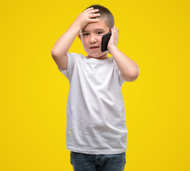 Dark haired little child using a smartphone stressed with hand on head, shocked with shame and surprise face, angry and frustrated. Fear and upset for mistake.