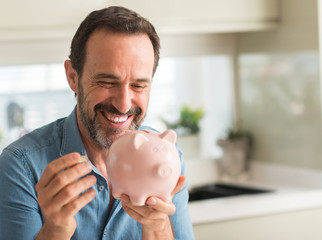 Middle age man save money on piggy bank with a happy face standing and smiling with a confident smile showing teeth