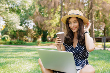 Adorable cute female student drinks coffee in summer green park while studying outside with laptop and smartphone. Student's lifestyle outside, college, university, back to study