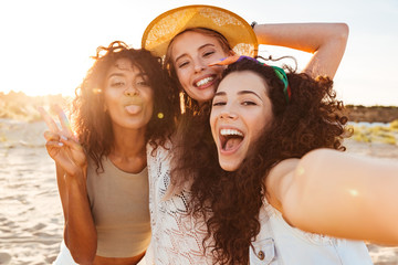 Obraz premium Three positive multiethnic women 20s in summer clothing smiling at camera, while taking selfie photo during holiday on nature