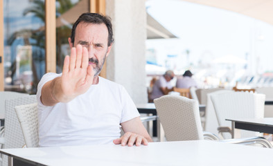 Handsome senior man sitting at terrace with open hand doing stop sign with serious and confident expression, defense gesture