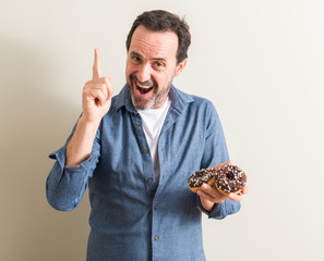 Senior man eating chocolate donut surprised with an idea or question pointing finger with happy...