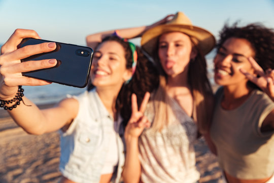 Blurry image of happy women 20s with different color of skin wearing summer clothing smiling and taking selfie photo on front-facing camera of smartphone, during walk at seaside