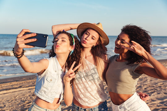 Three joyful beautiful women 20s with different color of skin wearing summer clothing showing victory sign, while taking selfie photo on smartphone with ocean background