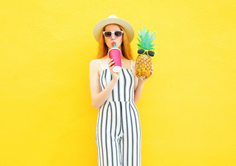 Fashion pretty woman with a pineapple, cup of juice in white striped pants, round hat on yellow background