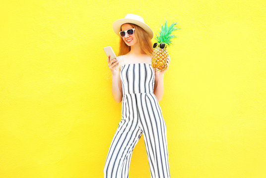 Fashion pretty woman holds a pineapple is using a smartphone in white striped pants, round hat on yellow background