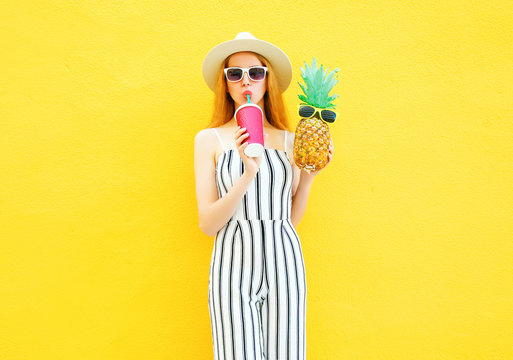 Fashion pretty woman with a pineapple, cup of juice in white striped pants, round hat on yellow background