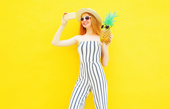 Fashion smiling woman and a pineapple is taking a picture on smartphone in white striped pants, round hat on yellow background