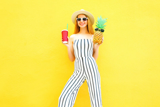 Fashion smiling woman holds a pineapple, cup of juice in white striped pants, round hat on yellow background