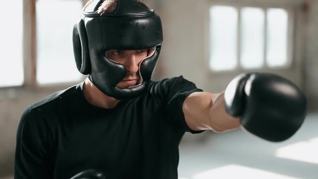 Aggressive boxer practises different punching techniques, wearing black boxing gloves and mask, having regular morning workout in large empty room