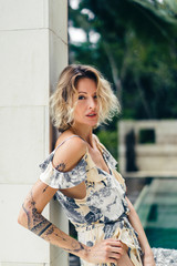 side view of attractive blond woman in dress standing at villa terrace with swimming pool on background