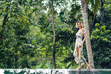 attractive blond woman in dress walking near swimming pool with green plants on background, ubud, bali, indonesia