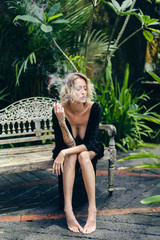 blond woman in black clothing smoking cigarette while resting on bench on terrace, ubud, bali, indonesia
