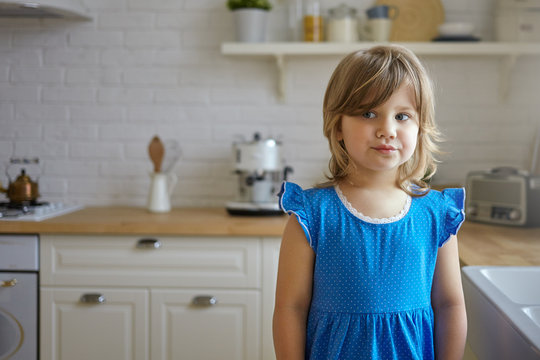 Indoor picture of cute funny little girl with chubby cheeks and fair hair posing in kitchen interior wearing blue dress looking sideways with playful facial expression. People and lifestyle