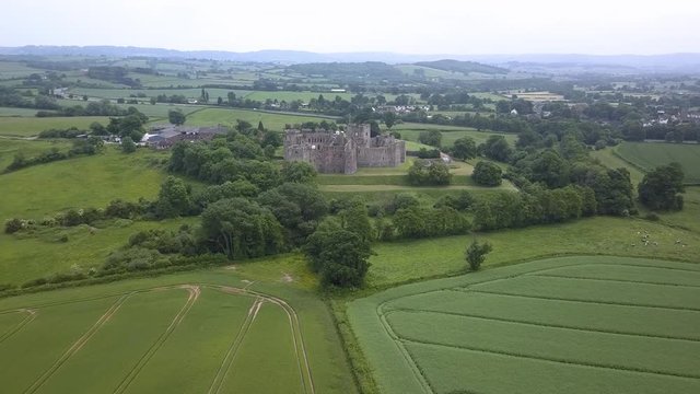 Aerial drone view flying towards the ruins of a medieval castle over green fields and farmland (Raglan Castle)
