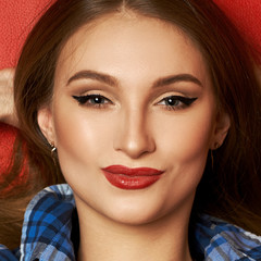 Portrait of cheerful beautiful caucasian woman dressed in blue checkered shirt smiling and posing with her hands behind head. Gorgeous long haired female model with red lips and black eye lines.