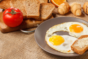 Plate with fried eggs, toast with butter, croissants, bread and tomato on wooden cutting board