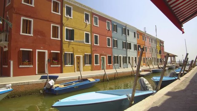 Walking along a calm river canal surrounded by picturesque colorful houses. Breathtaking shot of blue boats resting in front of different colored buildings in sunny Italy. Famous tourist attraction.