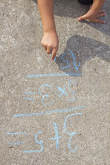 Girl kid thinking, writing and counting on mathematical equations with colored chalks on a pavement. School and vacation concept. Education concept. School and fun time.
