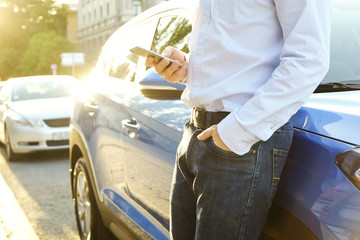 Young man in blue shirt formal ware leaning on his new caribbean blue car, using smartphone. Male holding blank screen cell phone in hands. Gps navigation concept. Close up, copy space, background.