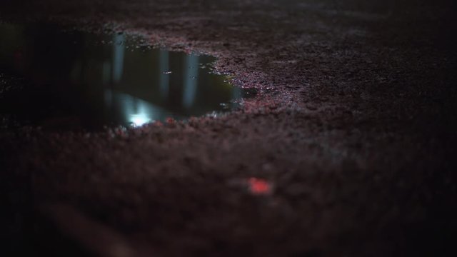 Closeup view of wet shining asphalt with reflection of electric city lights and raindrops in puddle at foggy humid night in slow motion