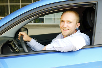 Young handsome businessman sitting behind steering wheel, test driving his new caribbean blue car. Smiling man in vehicle, satisfied with newely purchased automobile. Portrait, close up, background