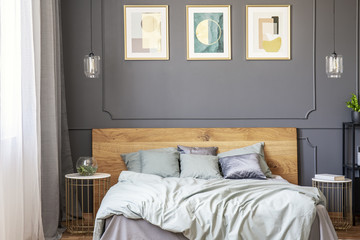 Posters on grey wall with molding above bed with wooden bedhead in bedroom interior. Real photo
