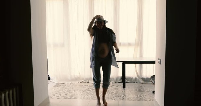 Young pregnant woman in hat and shirt having fun and dancing in room cheerfully