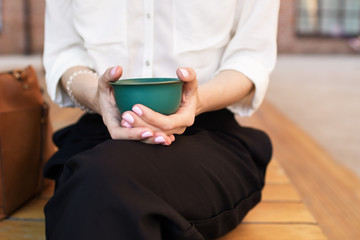 Woman holds an emerald bowl with matcha green tea in her hands over the green metallic table indoors. On the table there is a light bowl, jar, wooden stick and tea whisk. Closeup top view photo.