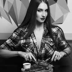 Gorgeous seductive woman dressed in unbuttoned checkered shirt sitting at table and eating delicious steak with knife and fork. Young beautiful female model at steakhouse or meat restaurant.