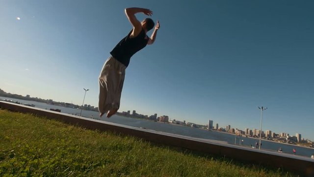 Young man professional acrobat performs a flip on a cliff overlooking the river and cityscape at sunset