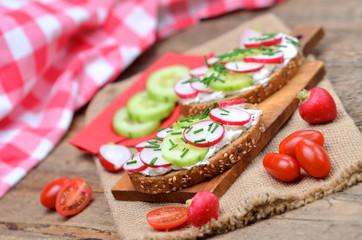 Wholemeal grain bread with curd cheese, fresh radish, cucumber and tomatoes on a wooden cutting board - concept of healthy fitness breakfast or snack
