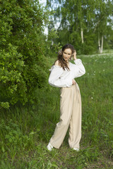 Beautiful stylish young girl wearing a trendy white shirt, beige trousers, and a pareo on her hair poses outdoors against a meadow and trees