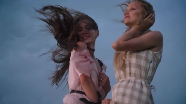 Two young charming girls in dresses is enjoing wind, touching hair in field in rainy day in summer, freedom concept, flirt concept, bottom view