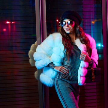 Pretty smiling female model wearing large round sunglasses, cap and white fur coat, posing against large glass window with shutter. Beautiful girl in trendy outerwear illuminated by pink lights.