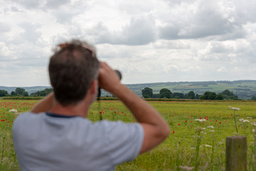 out of focus man in striped T shirt with beard looking at poppy field with binoculars